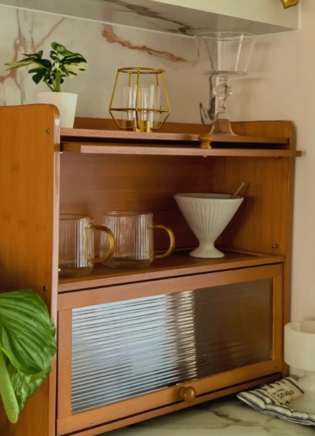 Bamboo cabinet on a kitchen counter with the top door lifted and slid in, showing glass mugs on the open shelf